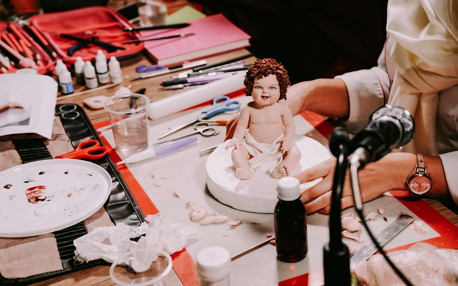 A handmade mixed-media art doll beside a notebook, ruler, archival folder, and camera on a clean worktable.